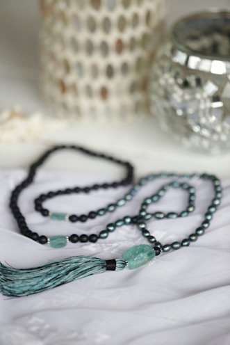 A beaded necklace with turquoise and black beads is elegantly arranged on a soft white fabric. The necklace features a prominent tassel and a larger turquoise bead. In the background, there are decorative items, including a metallic candle holder and a textured white ornament.