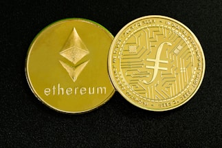 Two gold-colored coins are displayed on a dark surface. One coin features the Ethereum symbol and the word 'ethereum,' while the other has a circuit design and text around the edge.