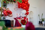 A festive setup featuring vibrant red balloons arranged in an arch above a table draped with burgundy cloth. A large white plush bear is placed next to the table, accompanied by various red and white decorations, including flowers and candles. Gold heart-shaped balloons add a touch of elegance, while lush greenery frames the scene. A golden birdcage and potted plants are positioned on the right, contributing to a lively atmosphere.