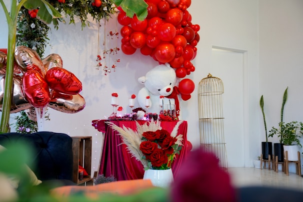 A festive setup featuring vibrant red balloons arranged in an arch above a table draped with burgundy cloth. A large white plush bear is placed next to the table, accompanied by various red and white decorations, including flowers and candles. Gold heart-shaped balloons add a touch of elegance, while lush greenery frames the scene. A golden birdcage and potted plants are positioned on the right, contributing to a lively atmosphere.