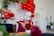 A festive setup featuring vibrant red balloons arranged in an arch above a table draped with burgundy cloth. A large white plush bear is placed next to the table, accompanied by various red and white decorations, including flowers and candles. Gold heart-shaped balloons add a touch of elegance, while lush greenery frames the scene. A golden birdcage and potted plants are positioned on the right, contributing to a lively atmosphere.
