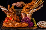 A variety of artisanal bread and baked goods are displayed on a dark surface. The centerpiece is a copper pot filled with an assortment of breadsticks, some of which are intricately shaped. Surrounding the pot are loaves of braided bread, vibrant purple bread slices, and rolls with red swirls. The warm tones and textures create a visually appealing assortment.