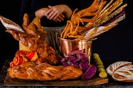 A variety of artisanal bread and baked goods are displayed on a dark surface. The centerpiece is a copper pot filled with an assortment of breadsticks, some of which are intricately shaped. Surrounding the pot are loaves of braided bread, vibrant purple bread slices, and rolls with red swirls. The warm tones and textures create a visually appealing assortment.