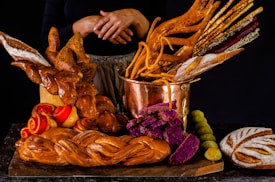 A variety of artisanal bread and baked goods are displayed on a dark surface. The centerpiece is a copper pot filled with an assortment of breadsticks, some of which are intricately shaped. Surrounding the pot are loaves of braided bread, vibrant purple bread slices, and rolls with red swirls. The warm tones and textures create a visually appealing assortment.