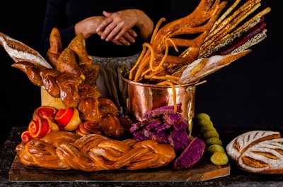 A variety of artisanal bread and baked goods are displayed on a dark surface. The centerpiece is a copper pot filled with an assortment of breadsticks, some of which are intricately shaped. Surrounding the pot are loaves of braided bread, vibrant purple bread slices, and rolls with red swirls. The warm tones and textures create a visually appealing assortment.