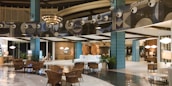 The image depicts a modern, well-lit hotel lobby with a spacious and elegant design. The area features stylish furniture, including wicker chairs and plush sofas, and polished flooring. Decorative elements include large blue-tiled pillars and artistic wall designs with circular patterns. The warm lighting and decorative plants add a cozy ambiance.