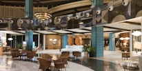 The image depicts a modern, well-lit hotel lobby with a spacious and elegant design. The area features stylish furniture, including wicker chairs and plush sofas, and polished flooring. Decorative elements include large blue-tiled pillars and artistic wall designs with circular patterns. The warm lighting and decorative plants add a cozy ambiance.