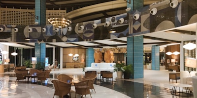 The image depicts a modern, well-lit hotel lobby with a spacious and elegant design. The area features stylish furniture, including wicker chairs and plush sofas, and polished flooring. Decorative elements include large blue-tiled pillars and artistic wall designs with circular patterns. The warm lighting and decorative plants add a cozy ambiance.