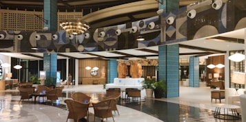 The image depicts a modern, well-lit hotel lobby with a spacious and elegant design. The area features stylish furniture, including wicker chairs and plush sofas, and polished flooring. Decorative elements include large blue-tiled pillars and artistic wall designs with circular patterns. The warm lighting and decorative plants add a cozy ambiance.