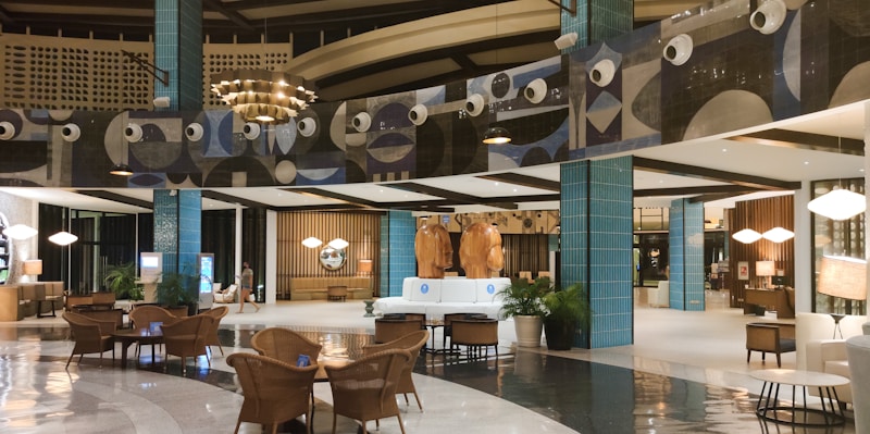 The image depicts a modern, well-lit hotel lobby with a spacious and elegant design. The area features stylish furniture, including wicker chairs and plush sofas, and polished flooring. Decorative elements include large blue-tiled pillars and artistic wall designs with circular patterns. The warm lighting and decorative plants add a cozy ambiance.
