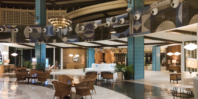 The image depicts a modern, well-lit hotel lobby with a spacious and elegant design. The area features stylish furniture, including wicker chairs and plush sofas, and polished flooring. Decorative elements include large blue-tiled pillars and artistic wall designs with circular patterns. The warm lighting and decorative plants add a cozy ambiance.