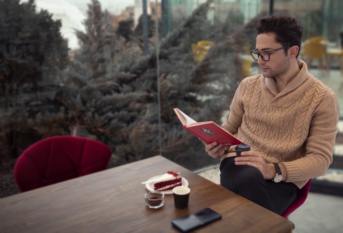 A young man at a cafe table reading a paperback in dim afternoon light, deep in concentration