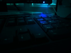 A close-up of a computer keyboard in dim blue-green lighting, highlighting several number keys and arrows. The background appears to include blurred, out-of-focus elements, suggesting a darkened room with electronic devices.