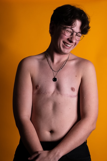 A person with short dark hair and glasses, smiling with eyes closed. They are shirtless, wearing a necklace, and standing against a warm yellow background.