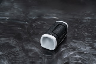 A portable speaker with a black and white exterior is placed on a wet surface. The object has water droplets on it, indicating exposure to moisture or water resistance. The background is dark and reflective.