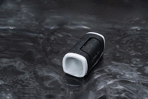 A portable speaker with a black and white exterior is placed on a wet surface. The object has water droplets on it, indicating exposure to moisture or water resistance. The background is dark and reflective.