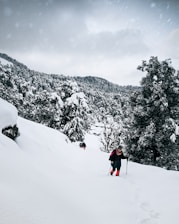 A person is hiking through a snowy landscape surrounded by heavily snow-laden trees. The sky is overcast with a hint of snowfall, and the trail is covered in deep snow. The hiker is using trekking poles and wearing bright red gaiters, which stand out against the white surroundings.