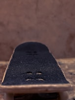 A close-up view of a skateboard deck with a textured black grip tape surface and two visible screws. The background is softly blurred in warm tones, emphasizing the detail in the skateboard deck.
