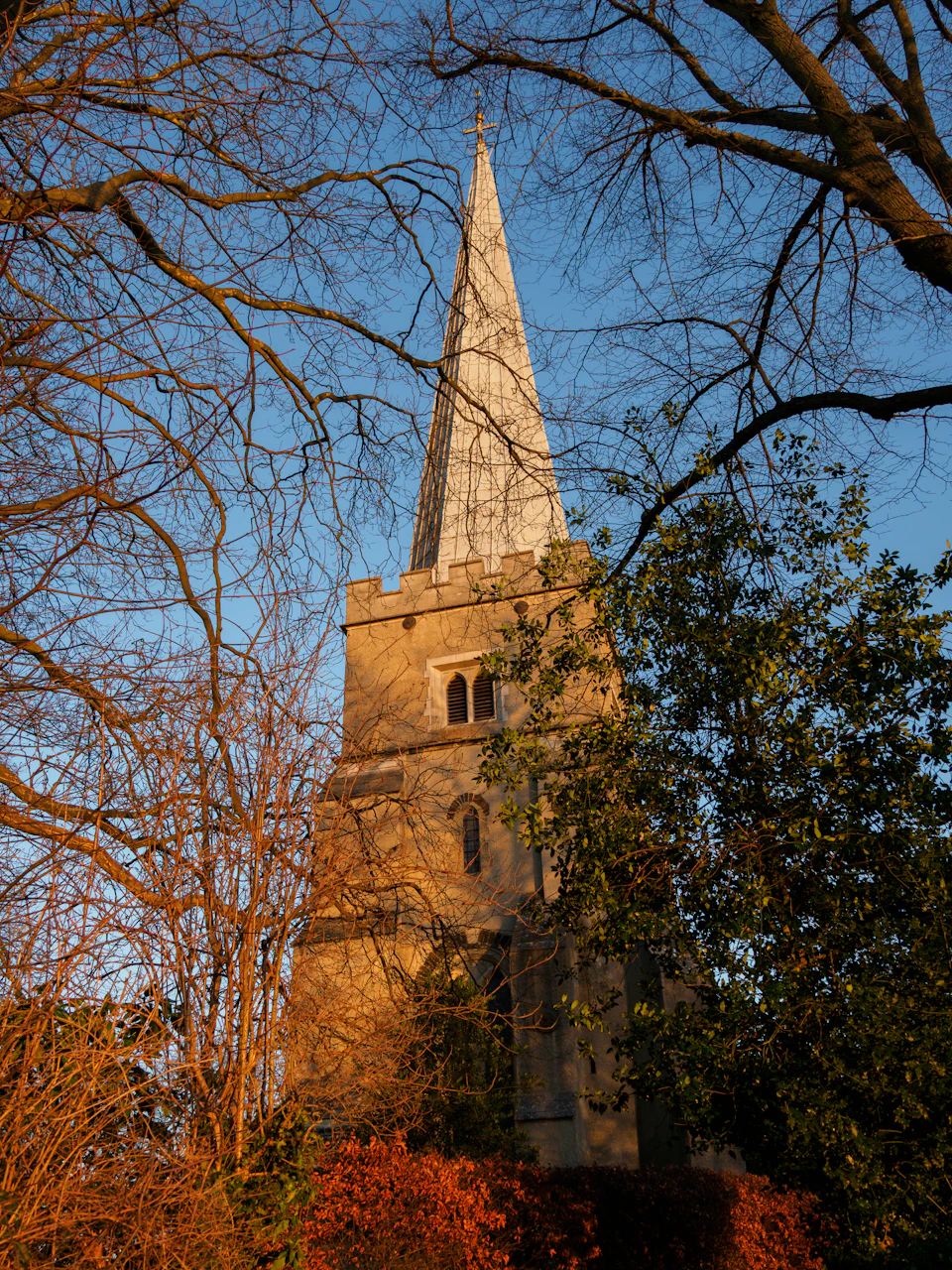 The image depicts a tall, white church with a pointed steeple and a cross at its pinnacle. The church is nestled amidst nature, surrounded by trees that have shed their leaves, creating a beautiful autumnal scene. The perspective of the photo is from below, looking up towards the church, emphasizing its height and grandeur against the backdrop of the sky.