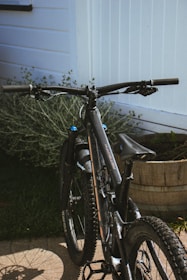 A mountain bike is parked on a patio next to a white wooden fence. The bike has thick tires suited for off-road cycling and features a sleek black frame. A patch of green grass and a wooden planter filled with plants are visible in the background.