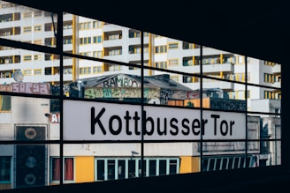 A cityscape view through large glass windows featuring a sign that says 'Kottbusser Tor'. The background includes tall, modern buildings with yellow window accents and various graffiti art. The scene is urban with elements of street culture and architecture.