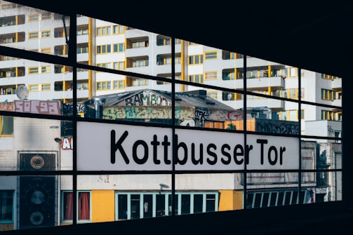 A cityscape view through large glass windows featuring a sign that says 'Kottbusser Tor'. The background includes tall, modern buildings with yellow window accents and various graffiti art. The scene is urban with elements of street culture and architecture.