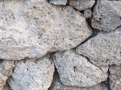 A close-up of a textured stone wall composed of rough, irregular limestone rocks fitting closely together. The surface shows various shades of gray and off-white, with visible pores and crevices.