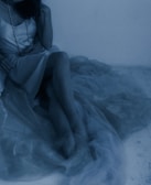 A person wearing a silky dress sits with legs crossed, partly draped in sheer fabric. A long necklace with pearls adds detail, and the scene has a soft, dreamy ambiance.