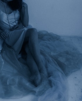 A person wearing a silky dress sits with legs crossed, partly draped in sheer fabric. A long necklace with pearls adds detail, and the scene has a soft, dreamy ambiance.