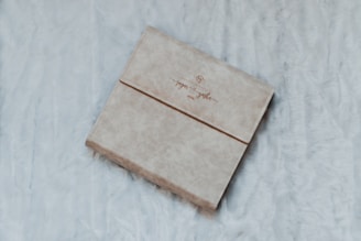A beige, textured cover book with elegant script on the front is placed on a soft, fluffy surface. The fabric appears cozy and inviting.