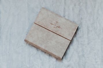 A beige, textured cover book with elegant script on the front is placed on a soft, fluffy surface. The fabric appears cozy and inviting.