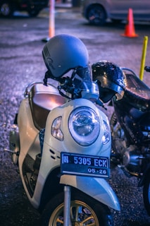 A close-up view of a parked motorbike with a gray helmet on its handle. The motorbike is wet, possibly from rain, and there are visible raindrops on the helmet. The background shows a blurred street with an orange cone and other vehicles.