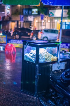 A street food cart is parked on a wet city street, illuminated by vibrant lights with a focus on a display case containing snacks like popcorn. The background features blurred vehicles and buildings, creating a lively urban atmosphere.