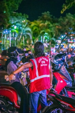 A person wearing a bright red reflective vest is interacting with a motorbike rider in a busy outdoor setting at night. The background is filled with lights and palm trees, creating a vibrant, illuminated atmosphere.
