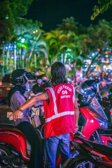 A person wearing a bright red reflective vest is interacting with a motorbike rider in a busy outdoor setting at night. The background is filled with lights and palm trees, creating a vibrant, illuminated atmosphere.