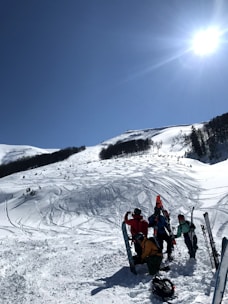 A group of people are gathered at the base of a snowy mountain slope, equipped with skis and snowboards. The sun is shining brightly in the clear blue sky, casting light on the untouched snow covering the landscape. Ski tracks are visible on the slope, suggesting previous activity. Trees are scattered around the mountainside.