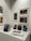 A gallery wall displaying various framed artworks featuring portraits and abstract compositions. Below the artworks, there is a table with a stack of books titled 'Pentimento Alexandre Farto aka Vhils' alongside several smaller prints and an open book displaying some images.