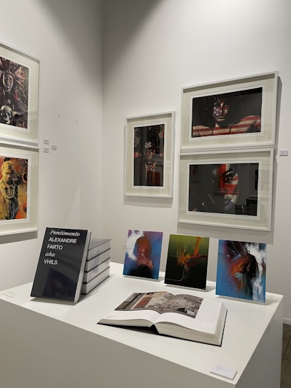 A gallery wall displaying various framed artworks featuring portraits and abstract compositions. Below the artworks, there is a table with a stack of books titled 'Pentimento Alexandre Farto aka Vhils' alongside several smaller prints and an open book displaying some images.