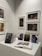 A gallery wall displaying various framed artworks featuring portraits and abstract compositions. Below the artworks, there is a table with a stack of books titled 'Pentimento Alexandre Farto aka Vhils' alongside several smaller prints and an open book displaying some images.