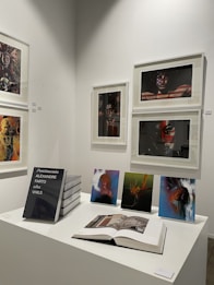 A gallery wall displaying various framed artworks featuring portraits and abstract compositions. Below the artworks, there is a table with a stack of books titled 'Pentimento Alexandre Farto aka Vhils' alongside several smaller prints and an open book displaying some images.