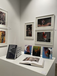 A gallery wall displaying various framed artworks featuring portraits and abstract compositions. Below the artworks, there is a table with a stack of books titled 'Pentimento Alexandre Farto aka Vhils' alongside several smaller prints and an open book displaying some images.