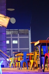A row of colorful metal chairs and tables is arranged on a wooden deck. The chairs feature vibrant shades of blue, yellow, red, and green. The setting is adjacent to a modern building with geometric windows, and the scene is illuminated by neon and ambient lighting, creating a modern urban ambiance.