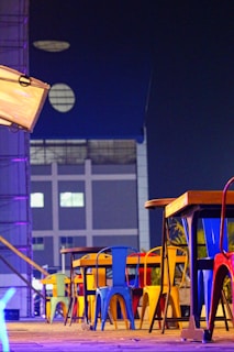 A row of colorful metal chairs and tables is arranged on a wooden deck. The chairs feature vibrant shades of blue, yellow, red, and green. The setting is adjacent to a modern building with geometric windows, and the scene is illuminated by neon and ambient lighting, creating a modern urban ambiance.