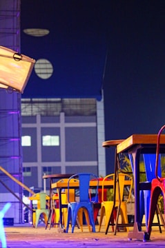 A row of colorful metal chairs and tables is arranged on a wooden deck. The chairs feature vibrant shades of blue, yellow, red, and green. The setting is adjacent to a modern building with geometric windows, and the scene is illuminated by neon and ambient lighting, creating a modern urban ambiance.
