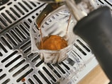 A rich, dark espresso shot being brewed into a clear glass measuring cup. The coffee machine's metal grate below the cup reflects the liquid’s deep brown color. The espresso has a creamy surface, indicating a well-extracted shot.