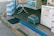 A sidewalk display at a Korean convenience store features neatly stacked packs of bottled water on the left. In front of the store's entrance, there's a blue plastic basket with an orange handle placed on a small stool. Signs in Korean promote the sale of various fruits and vegetables, along with a freezer for ice cream in the background.