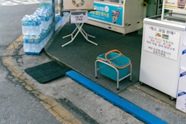 A sidewalk display at a Korean convenience store features neatly stacked packs of bottled water on the left. In front of the store's entrance, there's a blue plastic basket with an orange handle placed on a small stool. Signs in Korean promote the sale of various fruits and vegetables, along with a freezer for ice cream in the background.