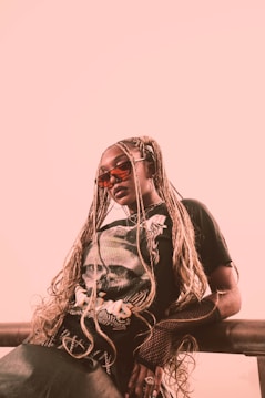 A person is leaning against a railing, wearing long braided hair, sunglasses, and a black graphic t-shirt. The background is a soft pink, and the overall look is edgy and stylish, with the outfit including fishnet gloves.