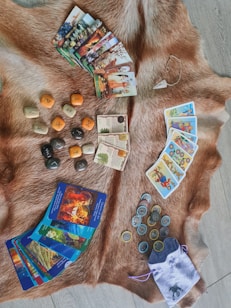 A variety of tarot and oracle cards are laid out on a brown fur rug, accompanied by a set of runes and coins. There's also a pendulum placed nearby. The items suggest a focus on mysticism and divination.