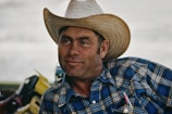 A person wearing a straw cowboy hat and a blue plaid shirt. The expression is relaxed and slightly smiling, with a pen visible in the shirt pocket. A blurred background shows parts of outdoor equipment or vehicles.